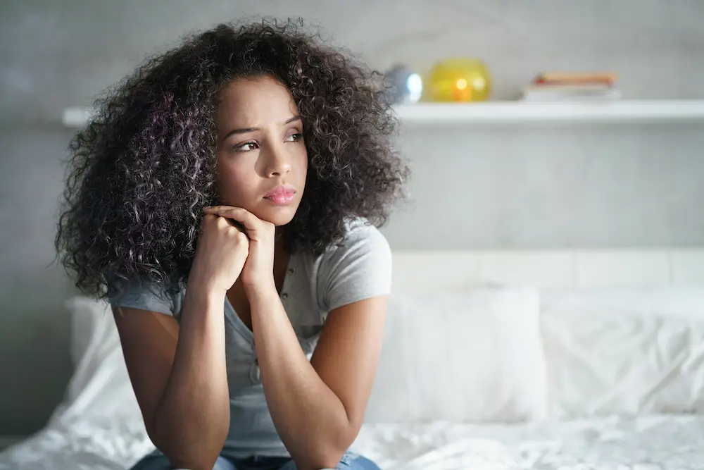 Young woman sitting on a bed resting her chin on her hands and looking thoughtful or worried